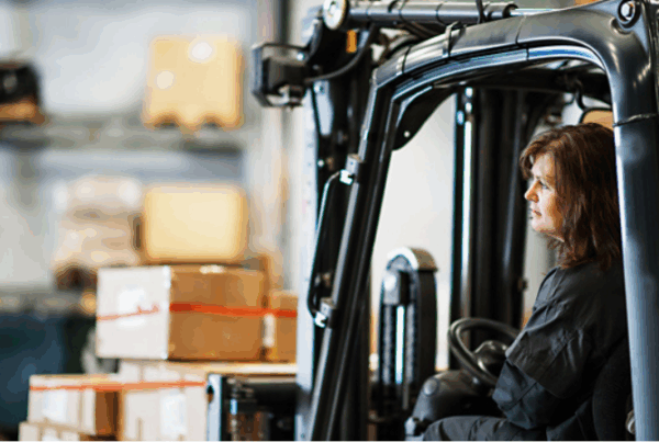 Kvinna i industriell miljö kör truck bland lastpallar – en symbol för begagnade maskiner och utrustning i aktiv användning.