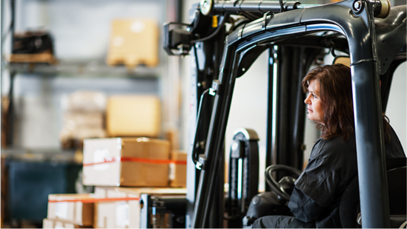 Kvinna i industriell miljö kör truck bland lastpallar – en symbol för begagnade maskiner och utrustning i aktiv användning.